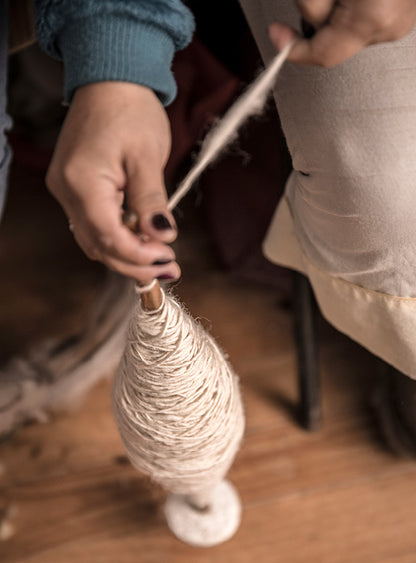 A close-up of a woman’s hand holding a skein of hand-spun wool, showcasing the texture and richness of the fibers. She is engaged in a crafting project, with colorful yarn and tools visible in the background.